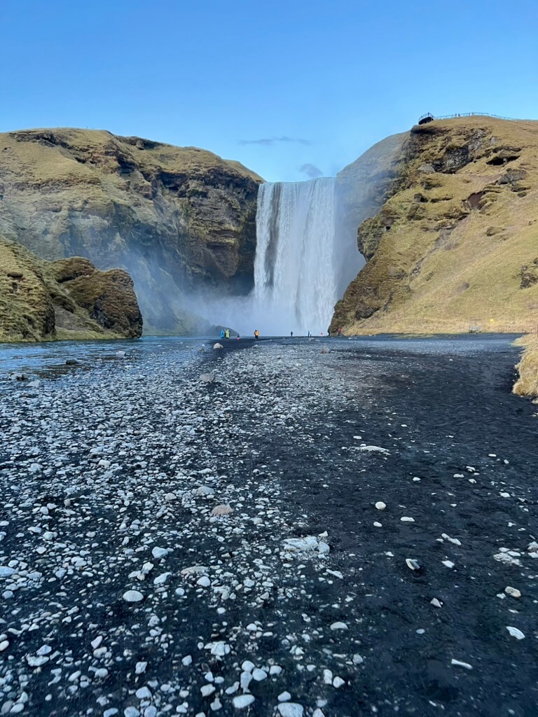 Tall waterfall cascading over rocky cliffs into a misty pool below. Dark volcanic rocks and pebbles line the foreground, with a clear blue sky above.