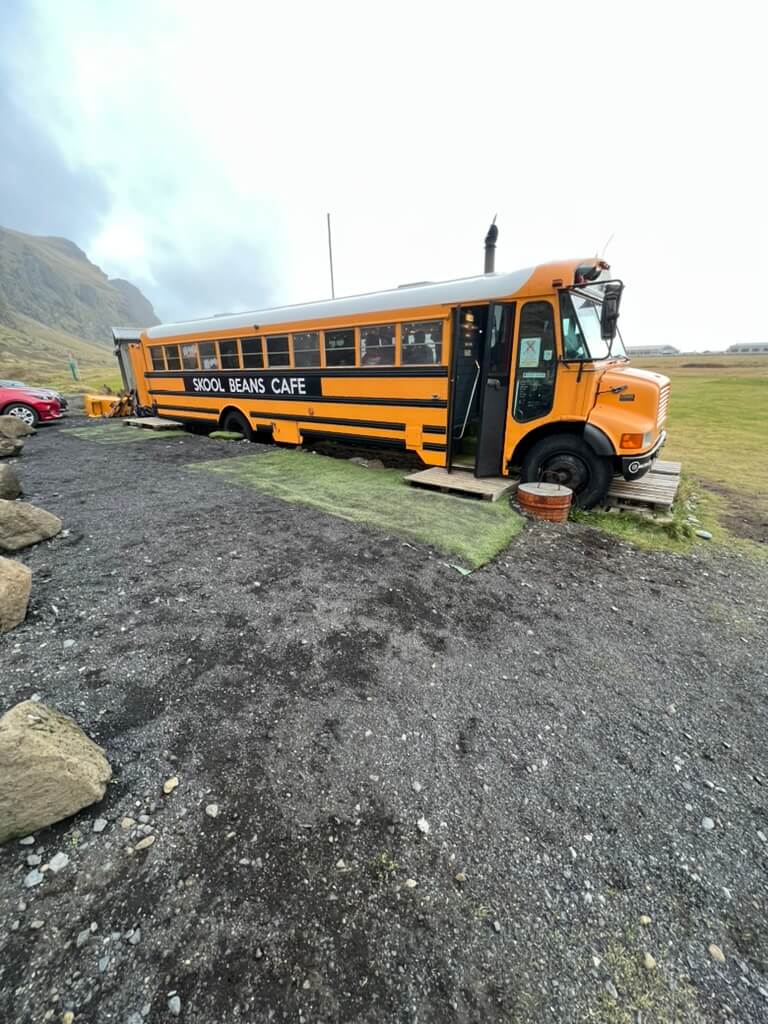 A yellow school bus converted into "Skool Beans Cafe" sits on a rural gravel lot with mountains in the background. A cozy, inviting atmosphere is conveyed.