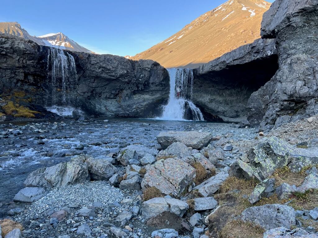 Mountain landscape with a serene waterfall cascading into a rocky stream, surrounded by rugged cliffs. Snow-capped peaks rise in the background under a clear blue sky.
