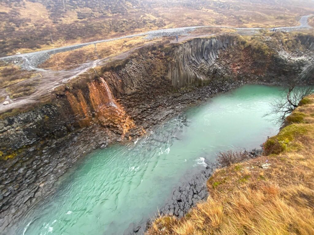 A small waterfall cascades over jagged, rocky cliffs into a turquoise pool. The surrounding landscape is rugged, with sparse brown vegetation.
