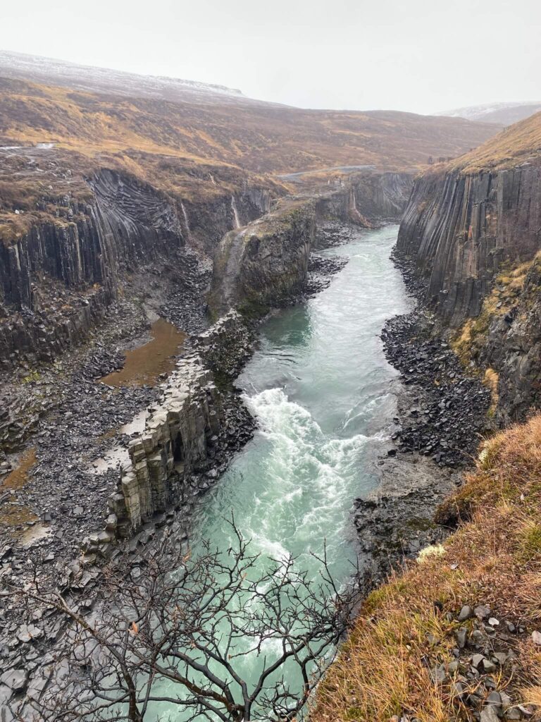 A turquoise river flows between rugged basalt cliffs under a gray, overcast sky. The landscape is barren and rocky, conveying a rugged, serene mood.