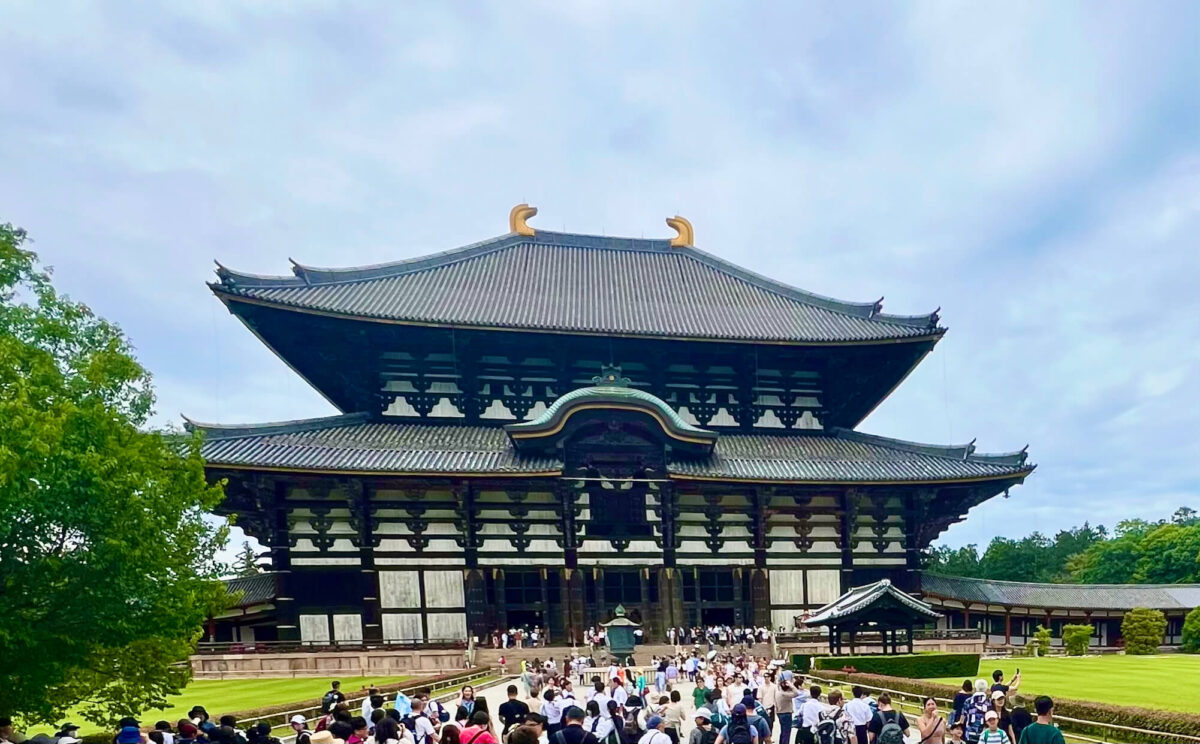 Large traditional Japanese temple building with a gray tiled roof under a cloudy sky. Crowds of people gather at the entrance, surrounded by lush greenery.