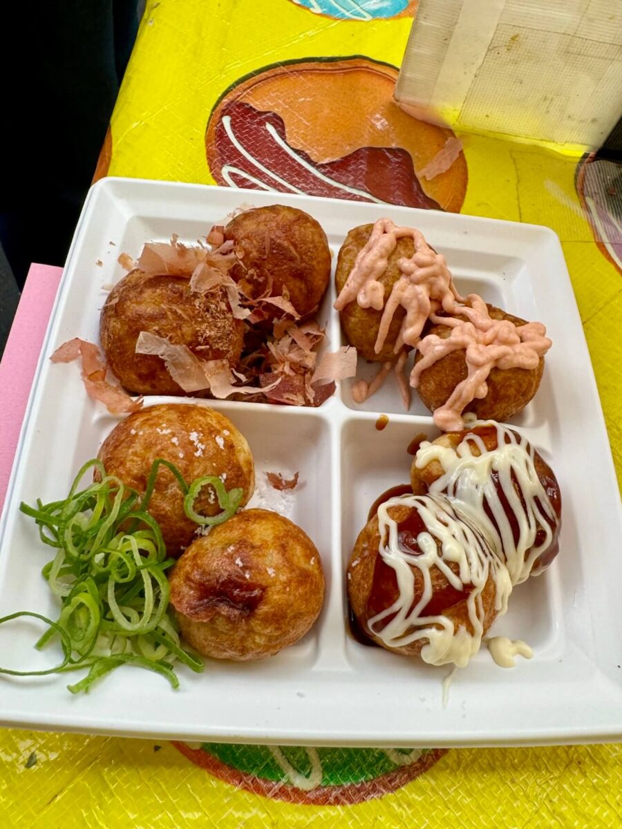 Four takoyaki varieties on a square white plate: topped with bonito flakes, mayo, pink sauce, and green onions, set on a colorful tablecloth.