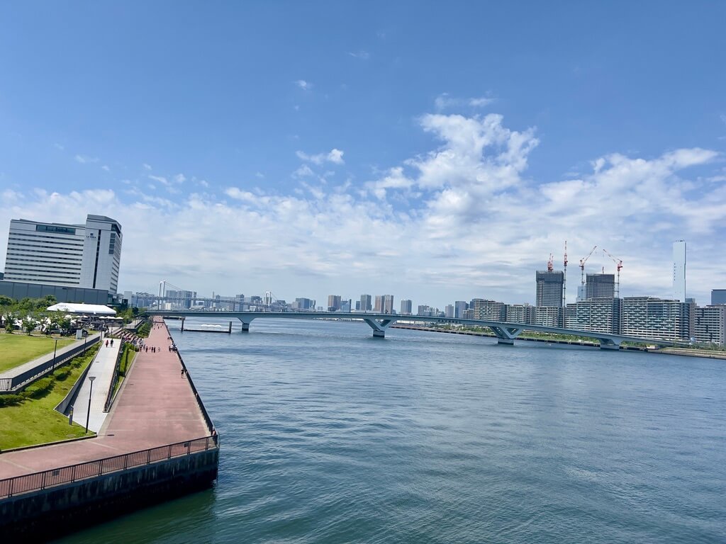 A scenic riverside view featuring a wide river under a blue sky, with a bridge and modern buildings, including construction cranes, in the background.