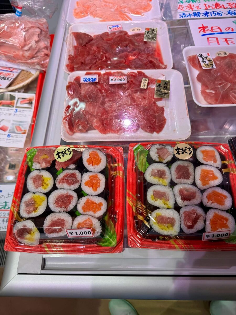 Assorted seafood and sushi products on display in a store. Fresh red fish slices are packaged in trays above sushi rolls in red containers, each priced at 1,000 yen.