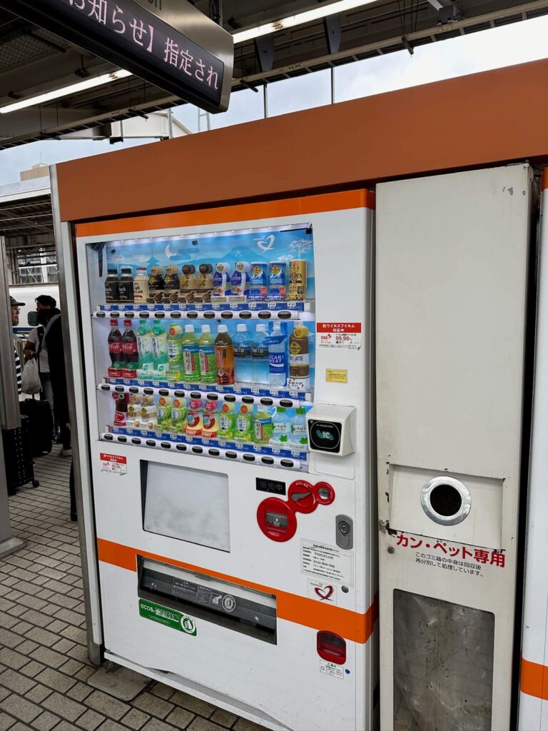 Vending machine at a train station offers a variety of drinks in bottles and cans. A recycling bin is attached, and signs are in Japanese. Bright lighting.