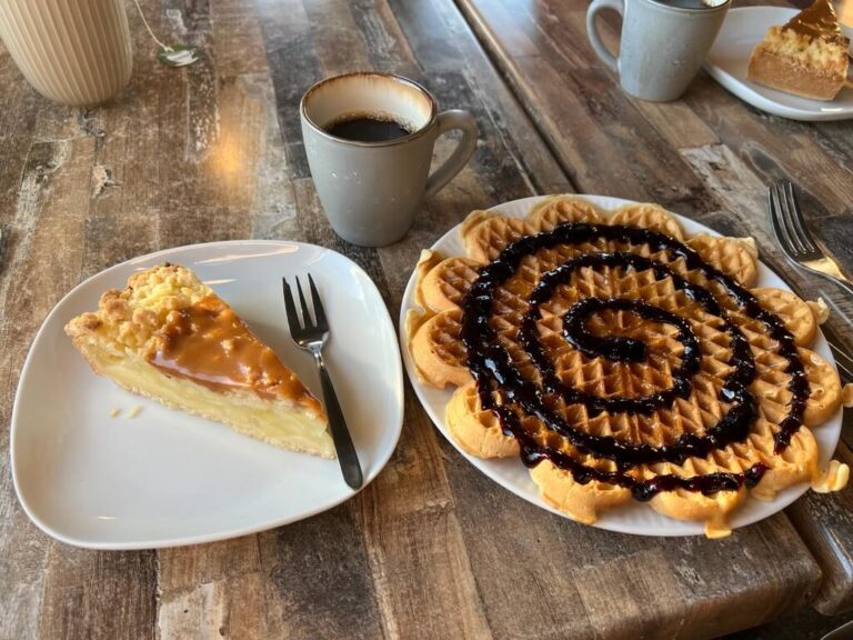 A slice of caramel-topped pie and a heart-shaped waffle with chocolate swirl sit on plates beside a cup of coffee on a rustic wooden table.