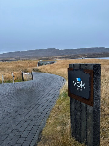 Curved stone path leads through golden grass to Vök Baths. The sky is overcast, creating a serene, tranquil atmosphere. Sign reads "Vök Baths."