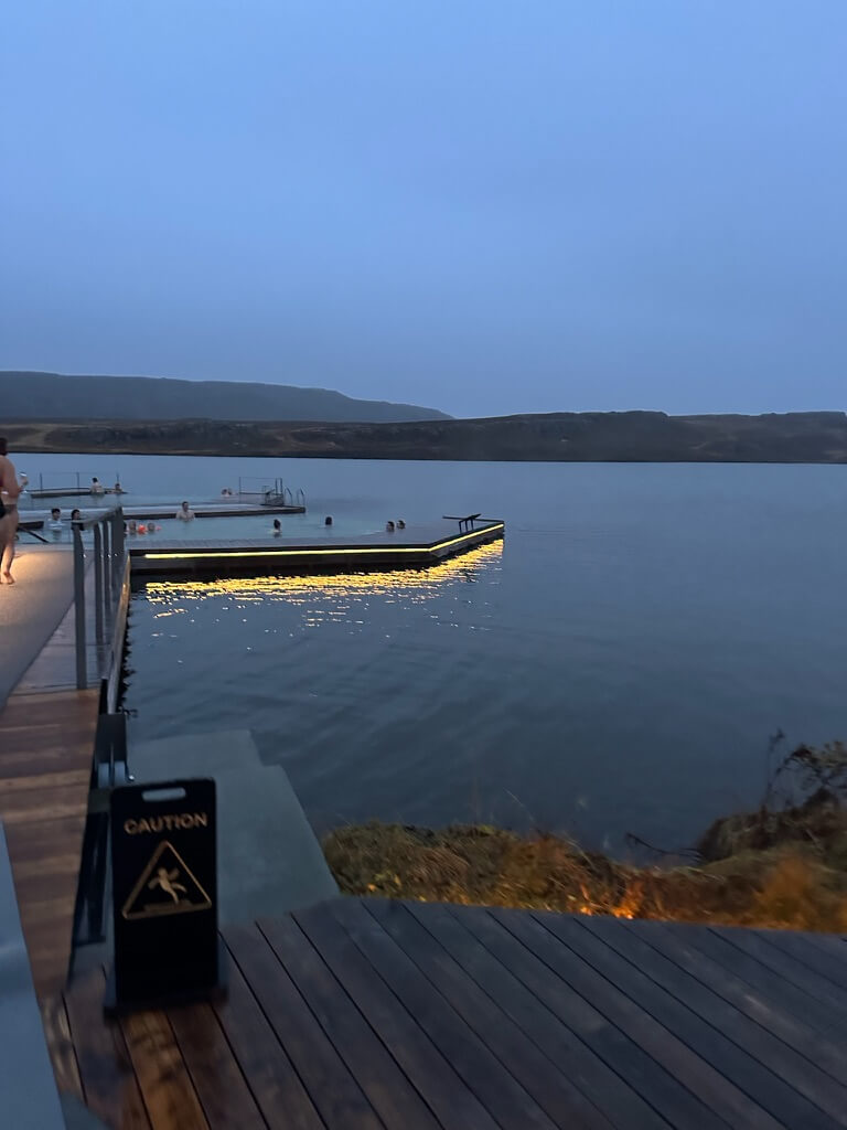 Evening scene of a geothermal pool by a calm lake with hills in the background. Soft lights illuminate the pool, creating a peaceful, serene atmosphere.