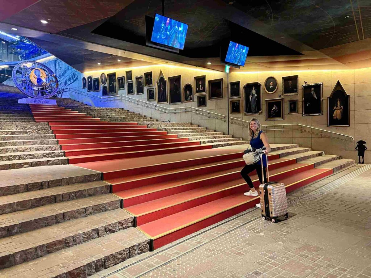 A woman with a suitcase stands on a red carpeted stairway, surrounded by framed portraits and displays, in a Harry Potter-themed setting. The atmosphere is magical and inviting.