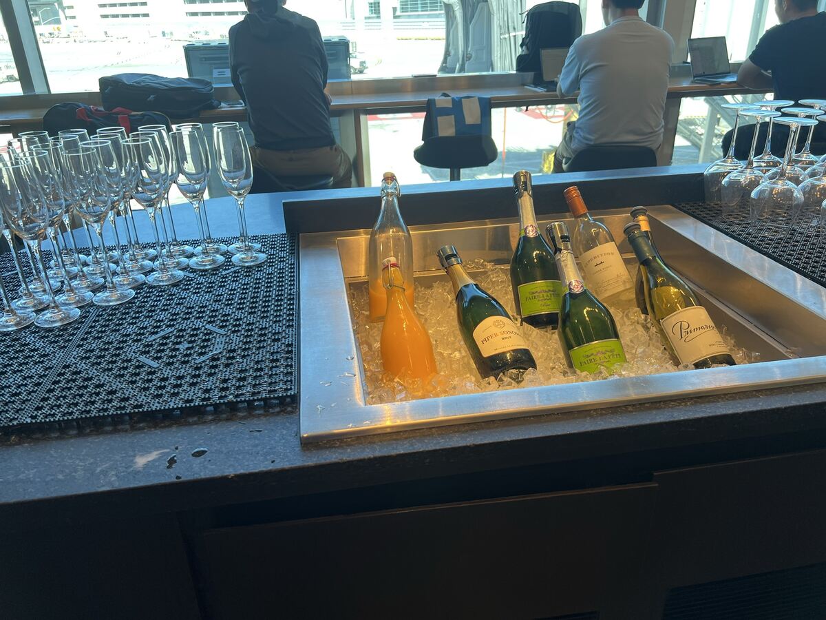 A collection of champagne bottles and orange juice on ice next to a row of empty glasses at a bar. Two people sit with laptops facing a window.