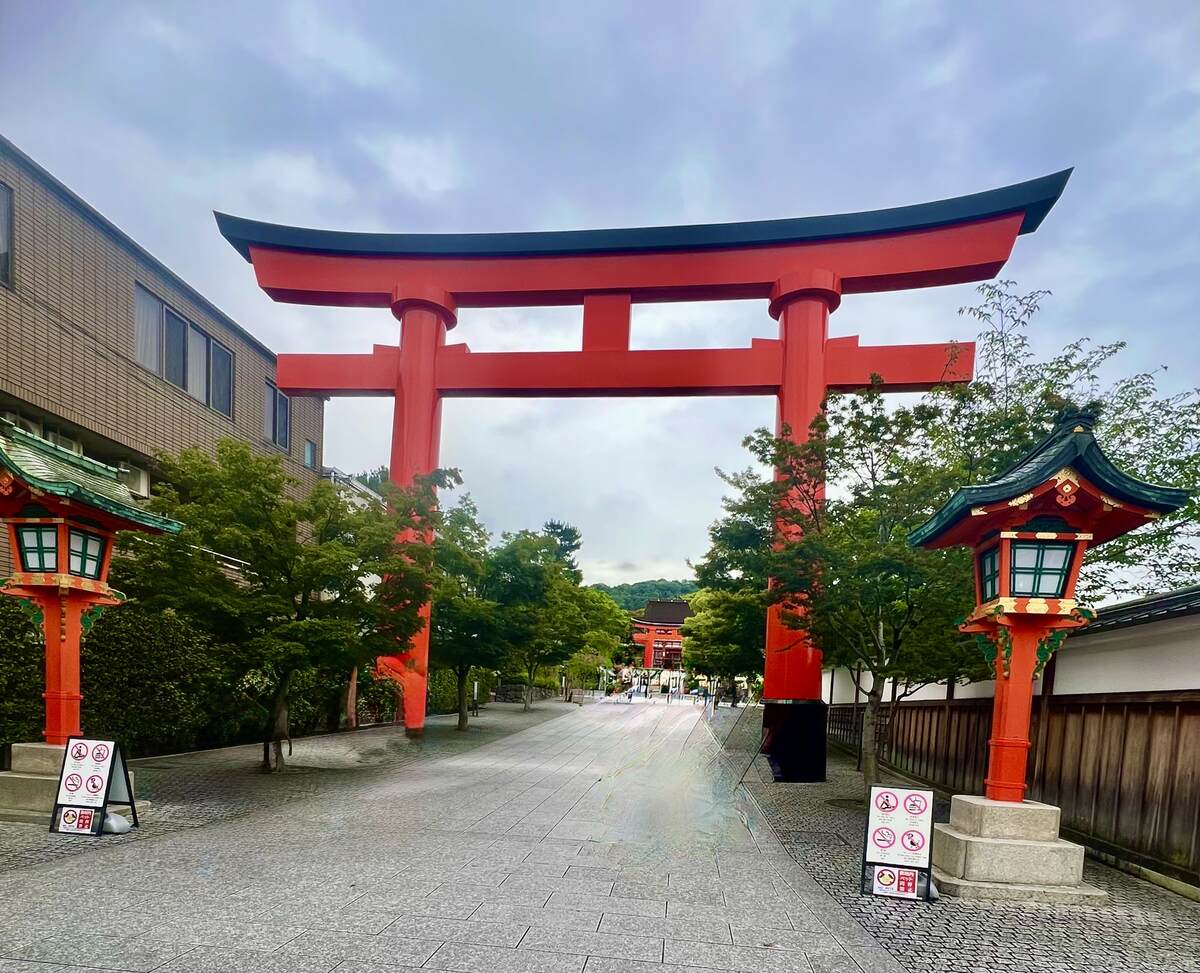 A large red torii gate stands at the entrance of a pathway lined with trees and traditional lanterns, under a cloudy sky, creating a serene and inviting atmosphere.