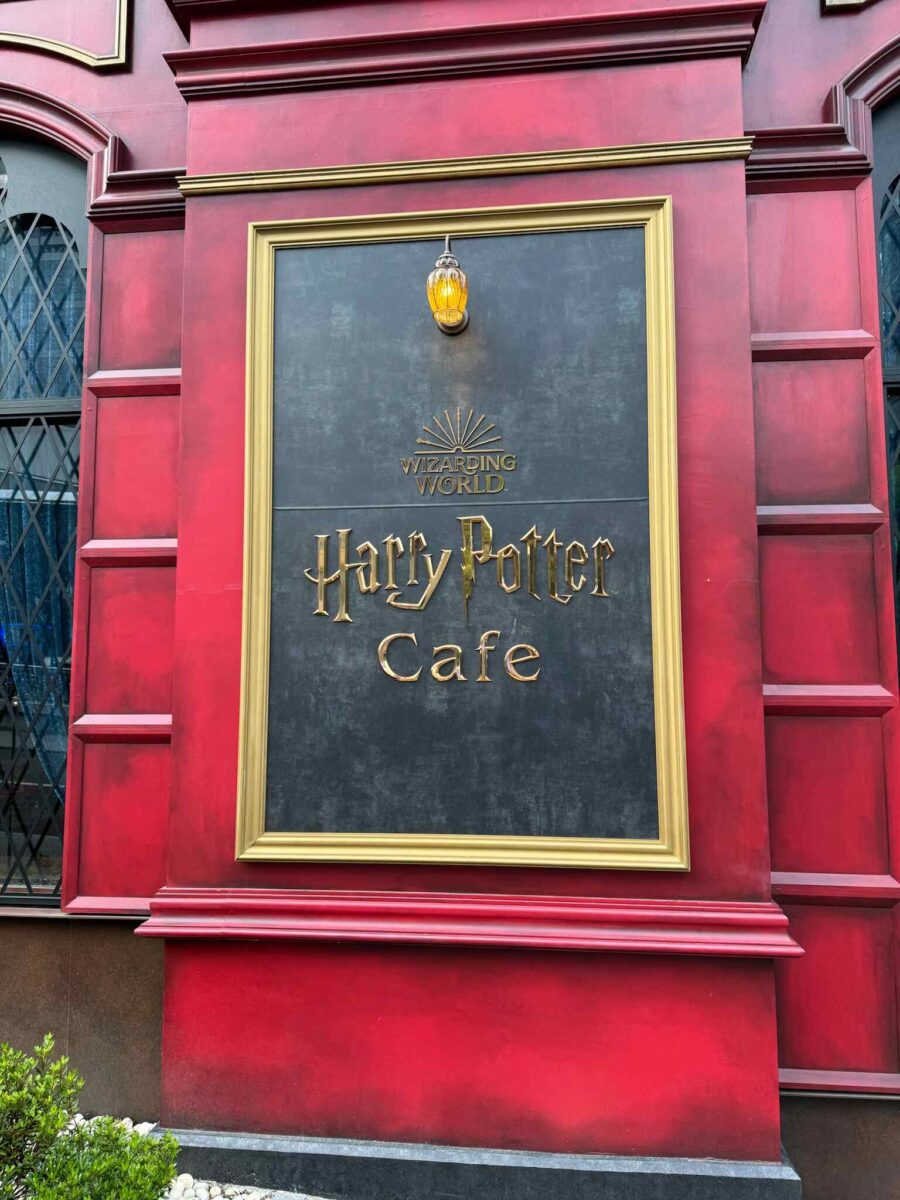 Signage for the Wizarding World "Harry Potter Cafe" on a red wall with gold trim. An ornate light fixture hangs above, suggesting a magical theme.