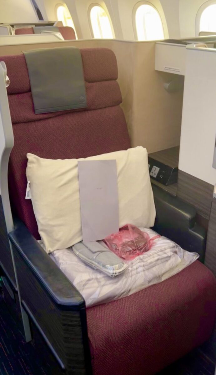 Luxurious airplane seat with a burgundy cushion, cream pillow, and a menu. Amenity kit and a blanket are neatly placed, evoking comfort and elegance.