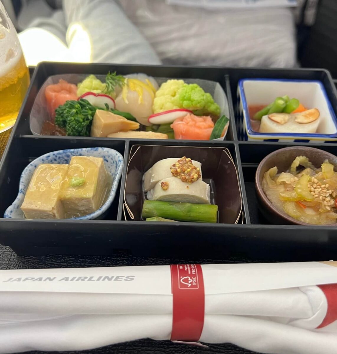 A Japan Airlines meal tray featuring assorted Japanese dishes, including tofu, fish, vegetables, and pickles, with chopsticks and a beverage nearby.
