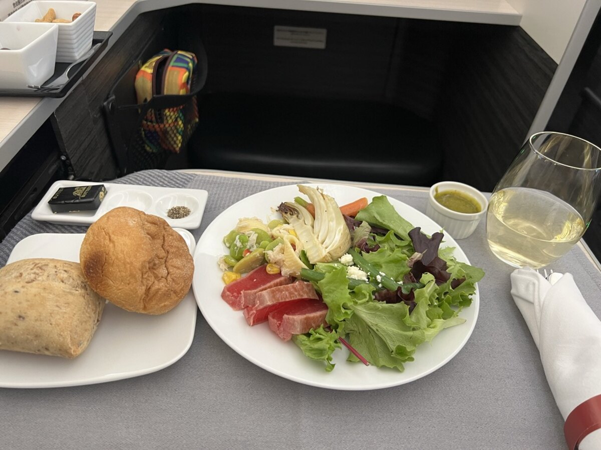 A plate of fresh salad with greens, tomatoes, and corn next to bread rolls. A glass of white wine and small condiment dishes are beside it on an airplane tray.