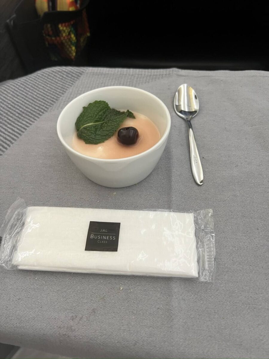A white bowl of panna cotta topped with a mint leaf and a cherry on a gray tablecloth in JAL Business Class, accompanied by a wrapped napkin and spoon.