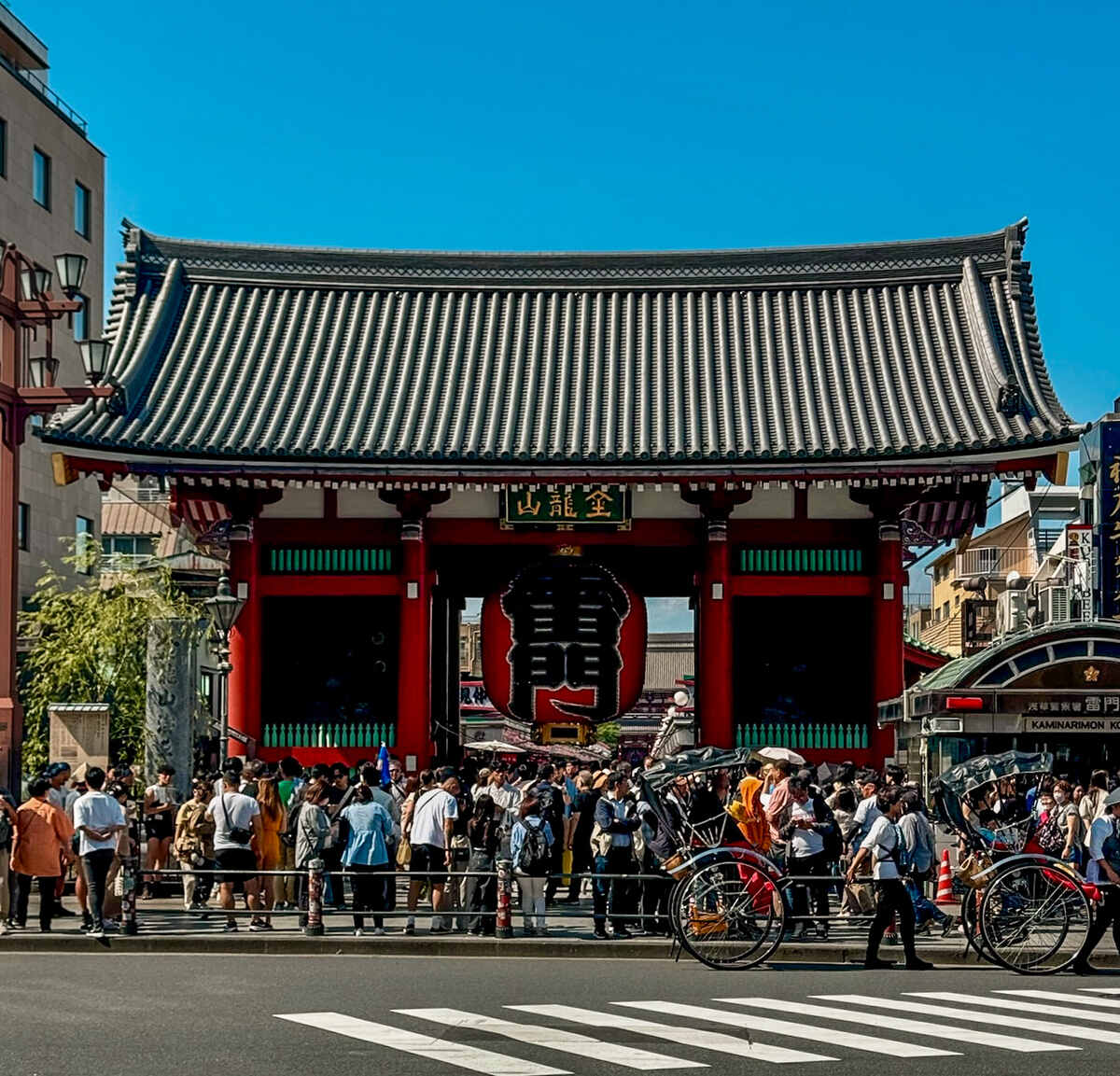 Crowded scene in front of a traditional Japanese gate, with a large red lantern and bustling tourists. Clear blue sky and vibrant, lively atmosphere.