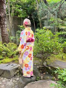 A person in a colorful floral kimono stands on a rock in a lush garden. They have a pink flower in their hair, surrounded by vibrant greenery.