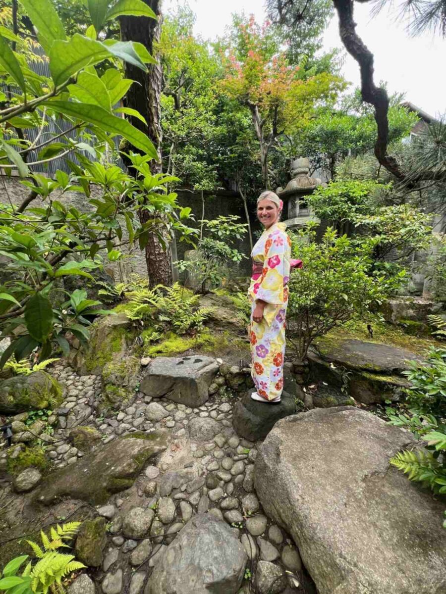 Woman in a floral kimono smiles while standing on a rock in a lush Japanese garden. Surrounding greenery, stone paths, and a calm, serene atmosphere.