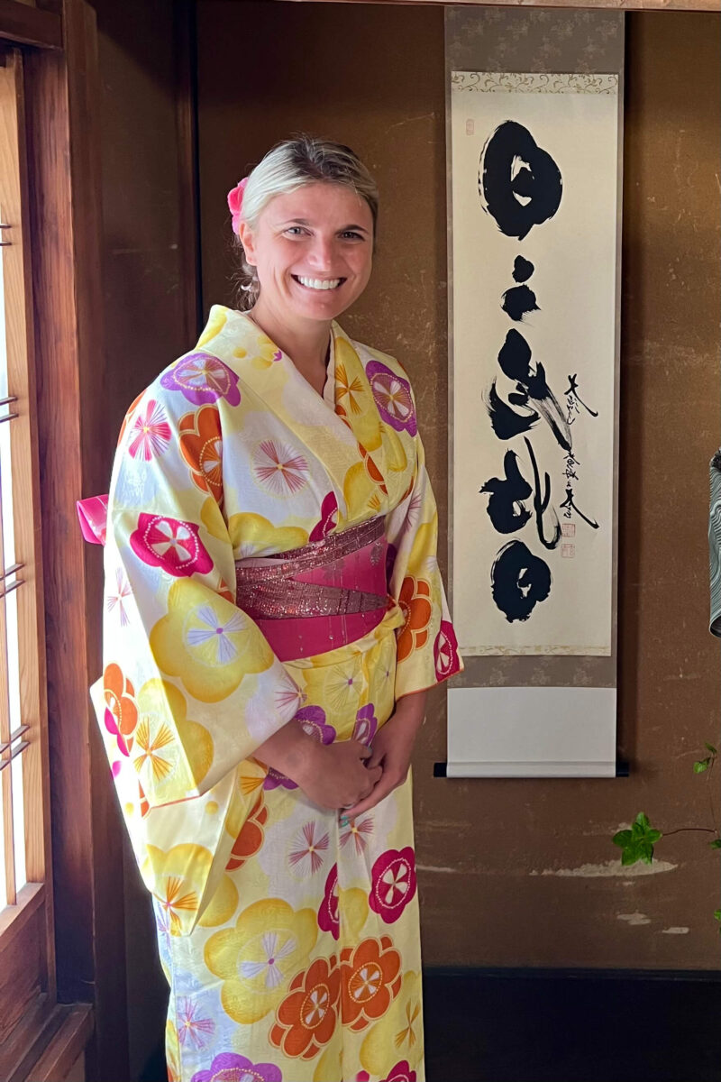 A smiling person in a colorful floral kimono stands beside a traditional Japanese scroll, conveying a warm, peaceful atmosphere in a softly lit room.