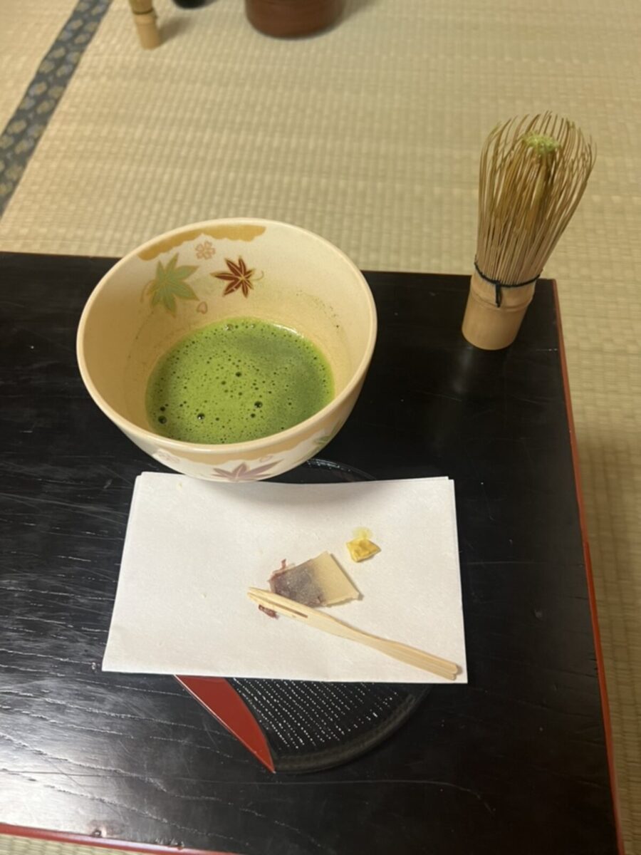 A bowl of frothy matcha green tea sits on a dark table, accompanied by a bamboo whisk and a small dessert on a napkin, creating a serene tea ceremony atmosphere.