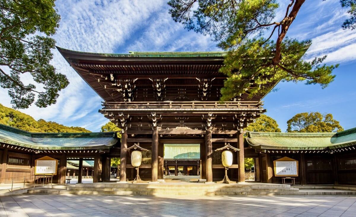 Wooden Shinto gate with curved roof at Meiji Shrine, Tokyo, surrounded by green trees under a blue sky, evoking serenity and tradition.