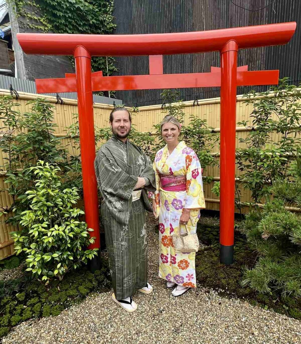 A man and woman stand in front of a red torii gate, dressed in traditional kimonos. The backdrop features lush greenery and a bamboo fence.