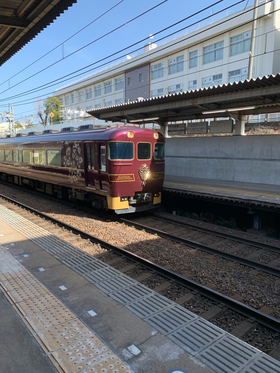 A maroon limited express train with floral designs sits on railway tracks at a platform. A modern building is visible in the background under a clear blue sky.