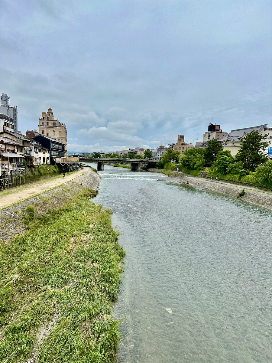 A serene river flows through a city, bordered by grassy banks and historic buildings. A bridge crosses over; the sky is cloudy, evoking calmness.