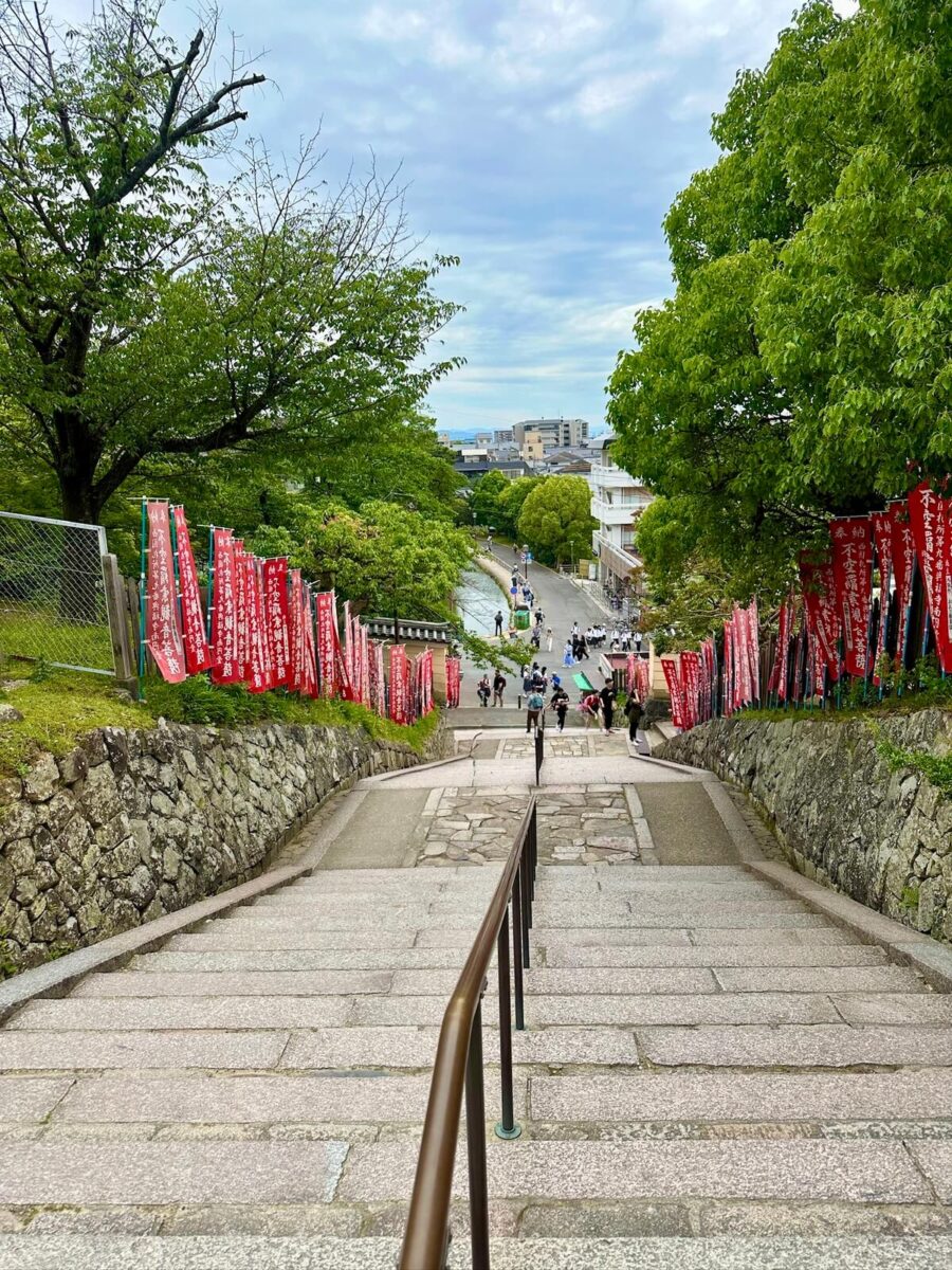 A stone staircase flanked by red banners and lush green trees descends towards a street. People are walking below, and a cloudy sky frames the scene.