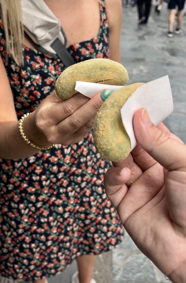 Two people holding Japanese mochi wrapped in paper. The sweets are sprinkled with green powder. Background shows a street scene with people.
