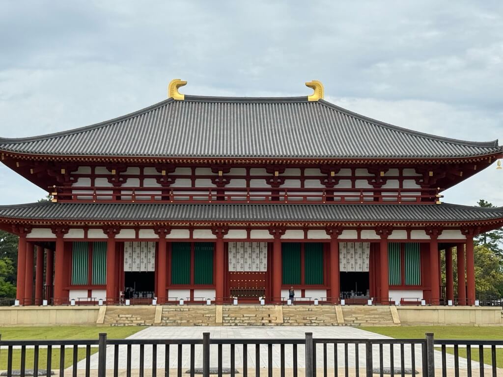 Traditional Japanese temple with red columns, gray tiled roof, and ornate golden accents against a cloudy sky. A peaceful, historic atmosphere.
