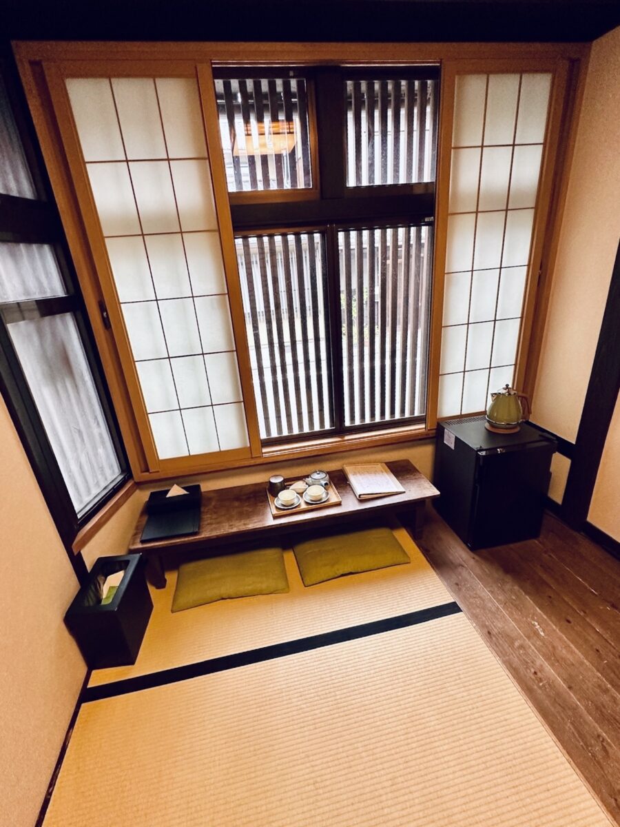 Traditional Japanese room with tatami mats, shoji screens, and low wooden table. Cushions on the floor and tea set create a serene ambiance.