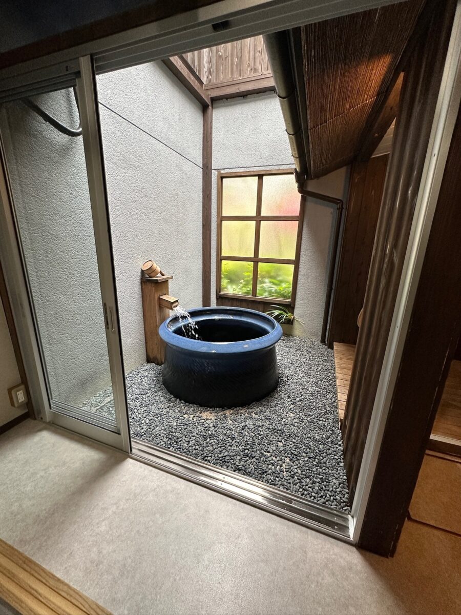 A serene Japanese indoor garden features a blue ceramic water basin with water gently pouring in. It's set on pebbles, framed by wooden walls and a window.