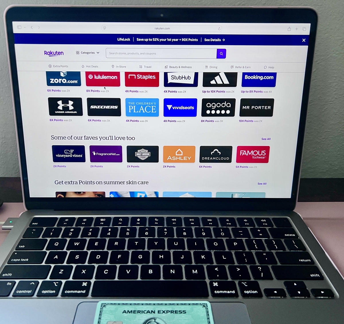 Laptop displaying a Rakuten webpage with logos of various brands like Zoro, Lululemon, and Staples. American Express card visible on the keyboard.