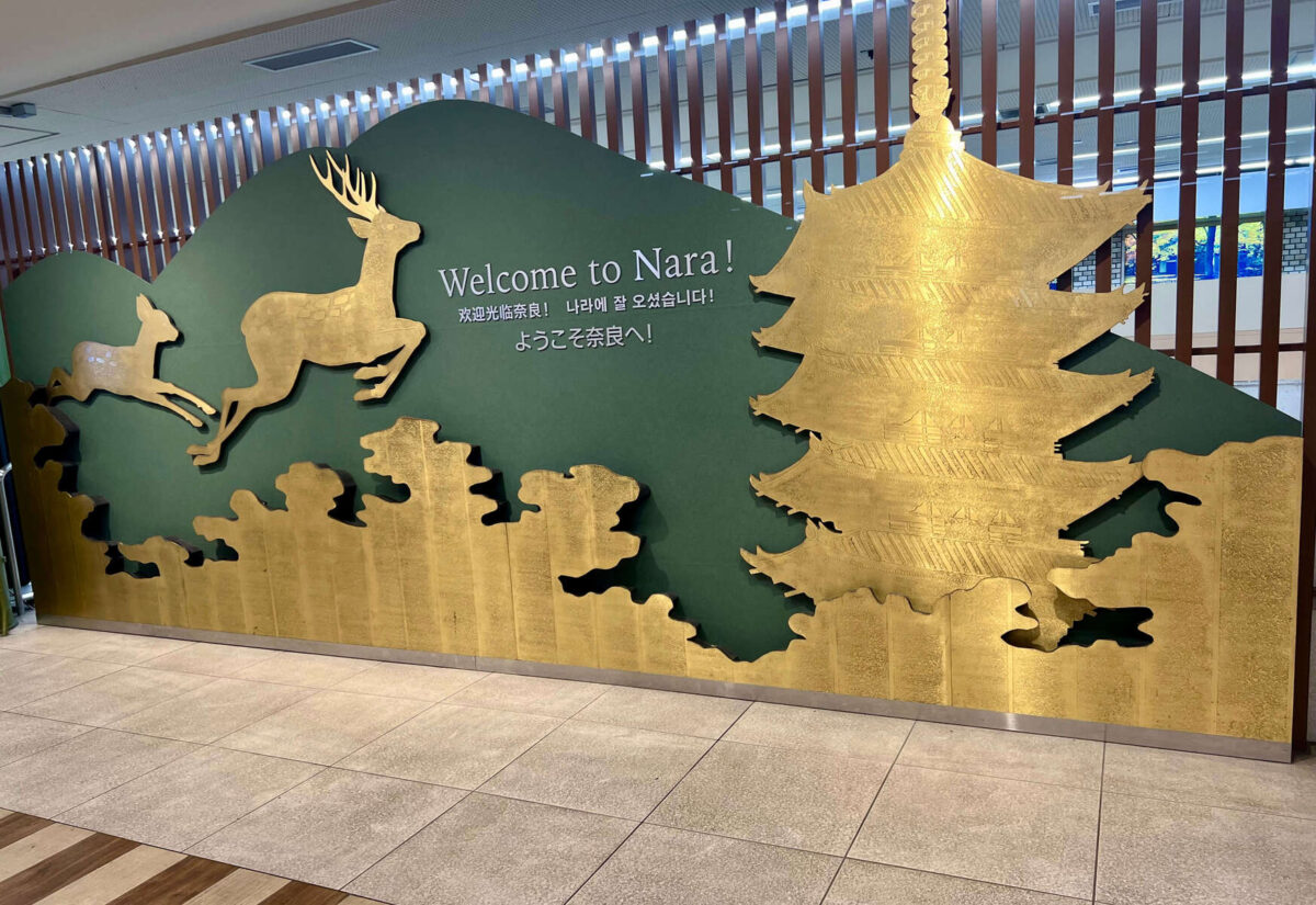 Golden silhouettes of a deer and pagoda against a green background, with "Welcome to Nara!" and greetings in multiple languages. Warm, inviting atmosphere.