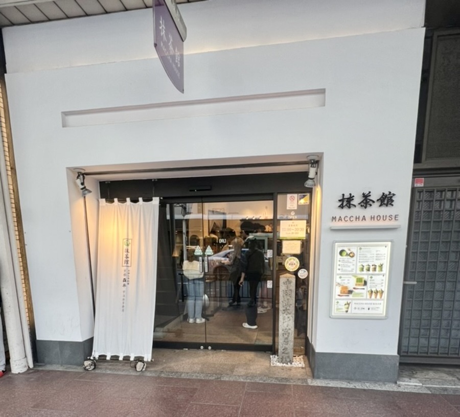 Entrance of Maccha House, featuring sliding glass doors with a white curtain on the left. A menu board is on the wall, with few people visible inside.
