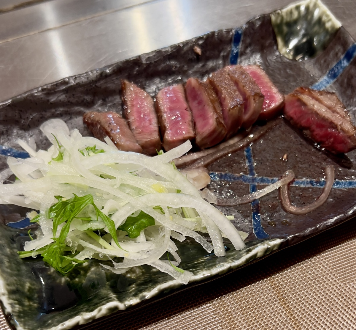 Slices of medium-rare A5 steak arranged on a ceramic plate beside a garnish of thinly sliced white onions and green leaves. The setting is elegant and appetizing.