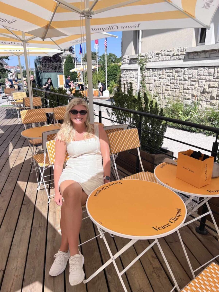 A woman in sunglasses and a white dress sits smiling on a patio. The bright yellow and white Veuve Clicquot-themed umbrellas and furniture create a cheerful, summery vibe.