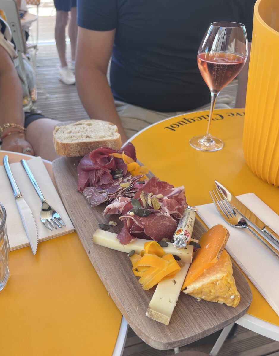 A colorful charcuterie board with assorted meats, cheeses, nuts, and bread on a wooden platter. It sits on a bright yellow table next to a glass of rosé wine and cutlery, conveying a relaxed, inviting atmosphere.
