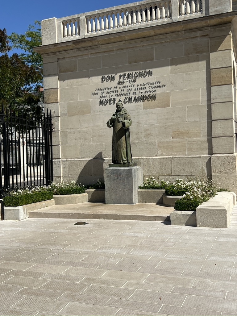 A statue of a monk holding a goblet stands on a pedestal against a stone wall. "Dom Perignon" and "Moet & Chandon" are inscribed above. Surrounded by greenery and a sunny courtyard.