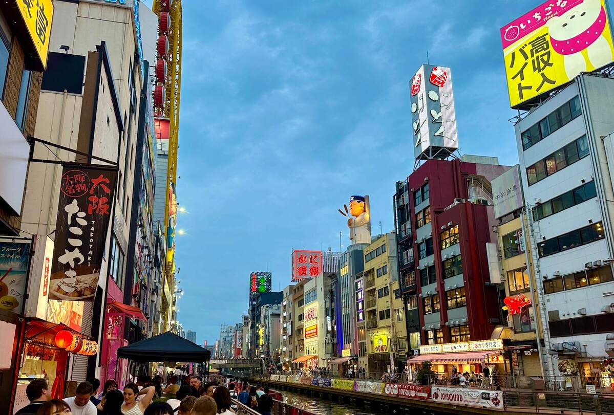 Bustling urban street in Osaka, Japan, with colorful neon signs and billboards. A lively crowd strolls along a canal under a vibrant evening sky.