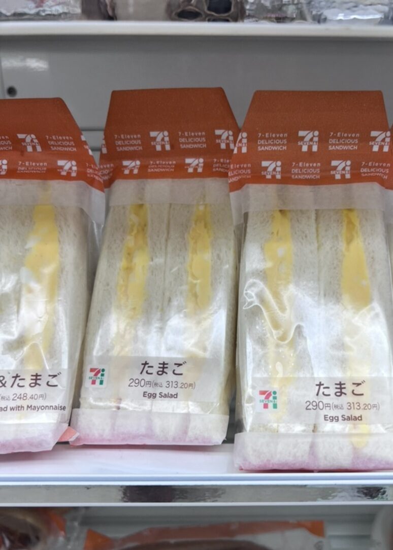 Packaged egg salad sandwiches on a store shelf, labeled in Japanese and English. The packaging features a 7-Eleven logo and orange accents.