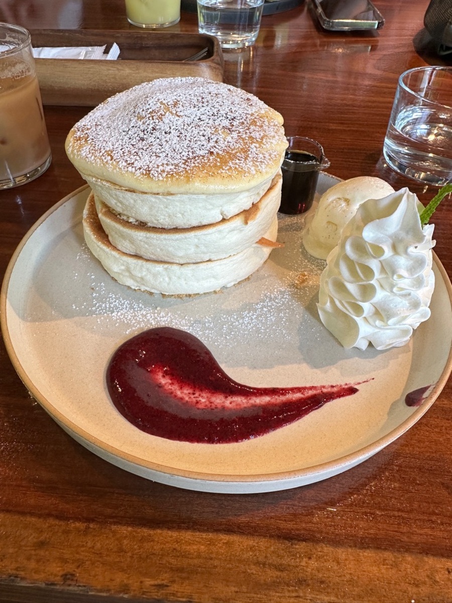 Fluffy pancakes dusted with powdered sugar are stacked high on a plate with whipped cream, a scoop of vanilla ice cream, and berry sauce.