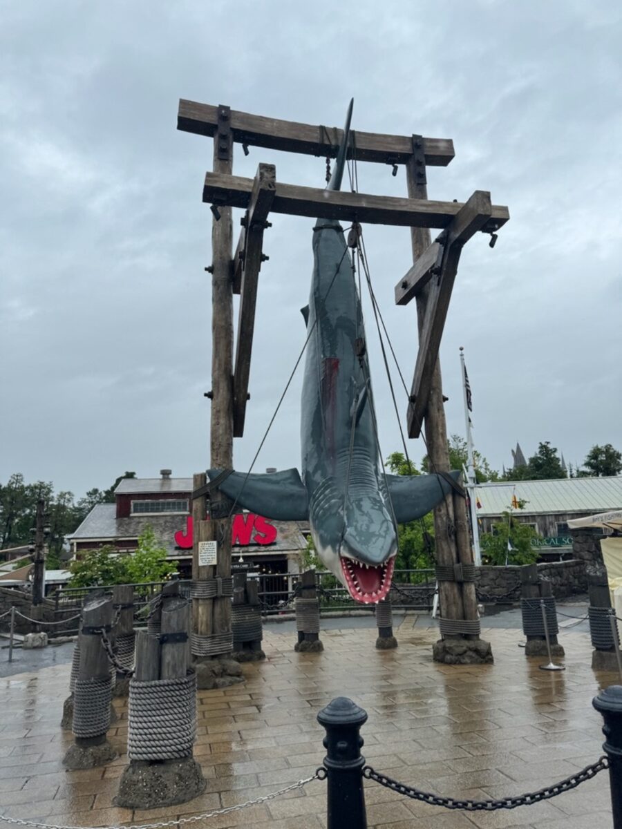 A large shark model is suspended vertically by ropes on a wooden structure resembling gallows. The ground is wet, suggesting recent rain. In the background, there's a "Jaws" sign, evoking a dramatic and cinematic tone.