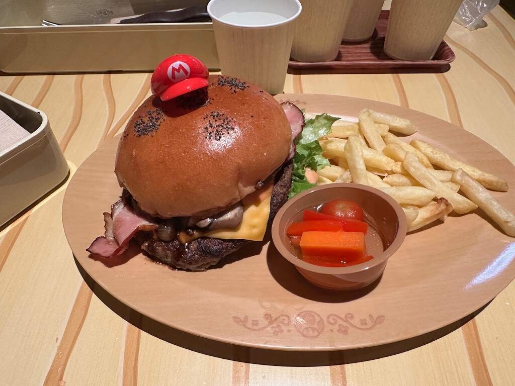 Burger with ham, cheese, lettuce, and a red Mario cap on top, served with fries and pickled vegetables on a wooden plate. Casual dining setting.