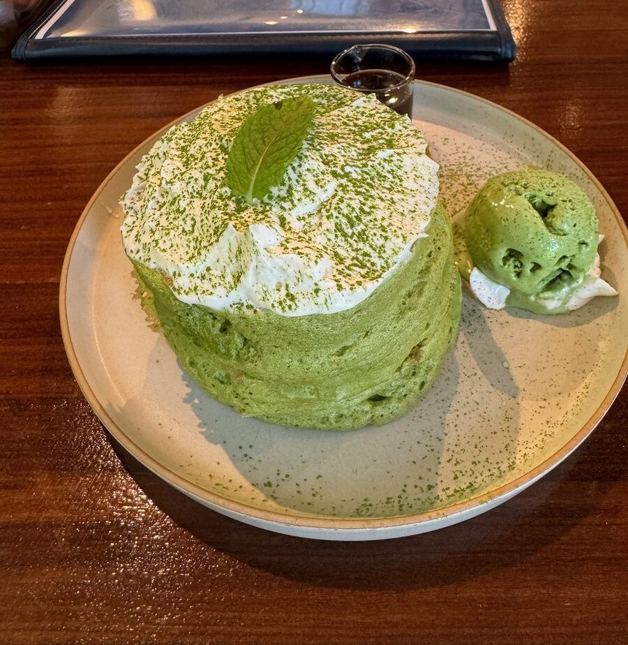 A plate of fluffy green matcha pancakes topped with whipped cream and a mint leaf, served with a scoop of green matcha ice cream and syrup, on a wooden table.