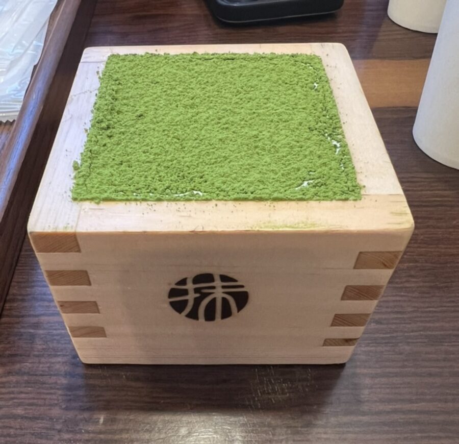 Wooden box with green matcha powder on top, featuring a decorative cut-out design on the side. Placed on a dark wooden table, conveying a calm, traditional feel.