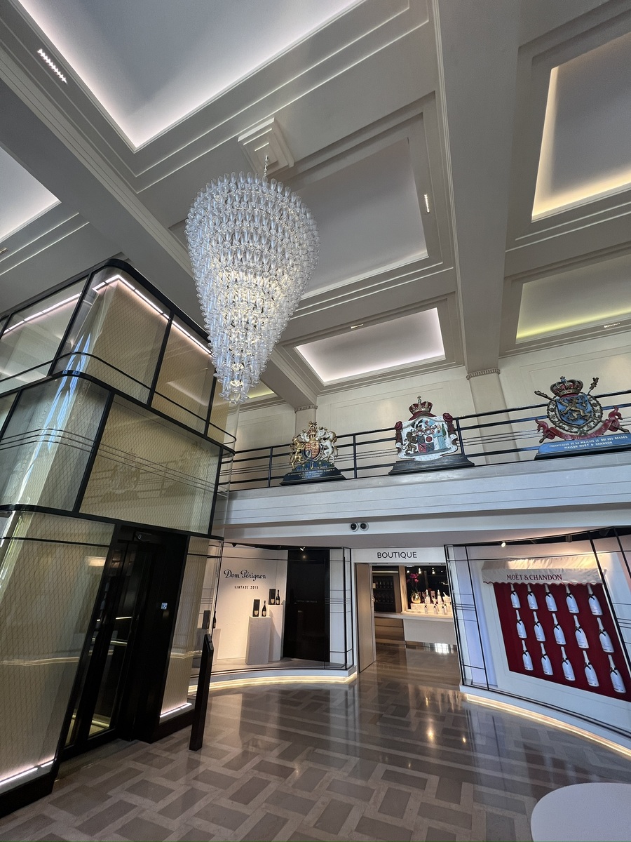 Luxurious lobby with a grand, cascading crystal chandelier, crests on the mezzanine, and boutiques displaying wine bottles. Bright, elegant ambiance.